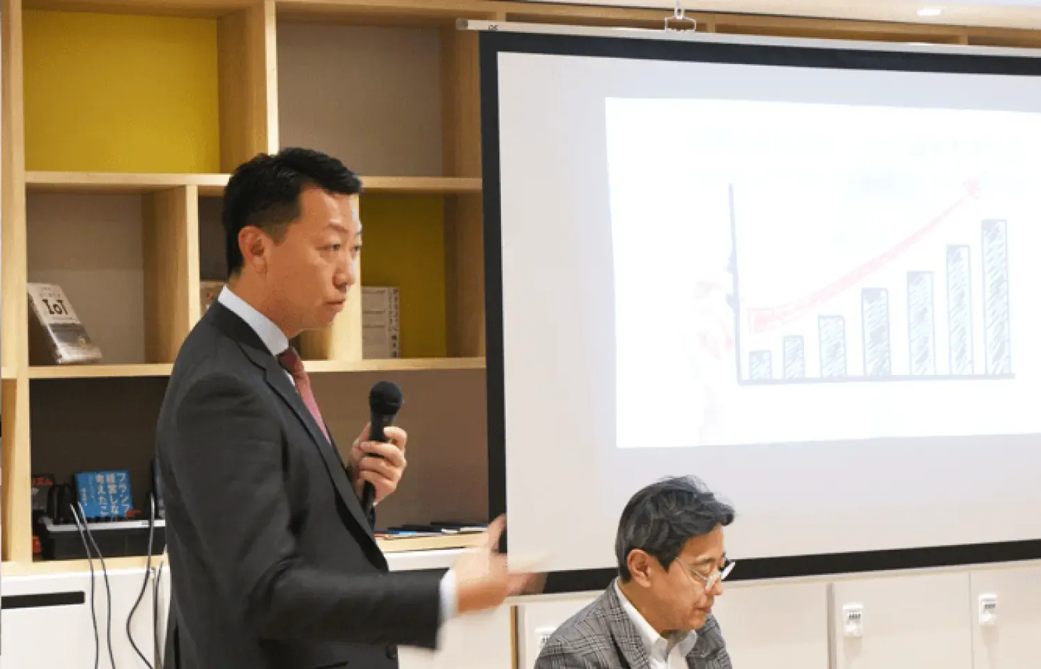 男性がマイクを持ち、会議で発表している様子。